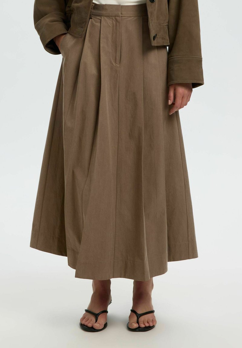 Femme portant une longue jupe plissée taupe associée à des sandales noires à brides et une veste taupe assortie sur un haut blanc.