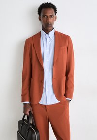 Blazer couleur rouille avec un pantalon assorti, tissu texturé, et une chemise rayée bleu clair en dessous. Sac à main noir avec double anse.