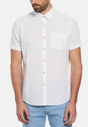 Chemise blanche à manches courtes avec un poche poitrine, texture légère et boutons marron clair. Portée avec un short bleu.