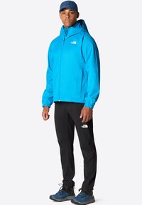 Modrá venkovní bunda s kapucí, elastickými manžetami a zipem vpředu, spárovaná s černými kalhotami a modrými turistickými botami. Výrobky mají branding.