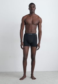 Fekete pamut boxer alsónadrág, elasztikus derékpánttal, amelyen a Calvin Klein logó látható, férfi alak viseli, semleges háttér előtt.