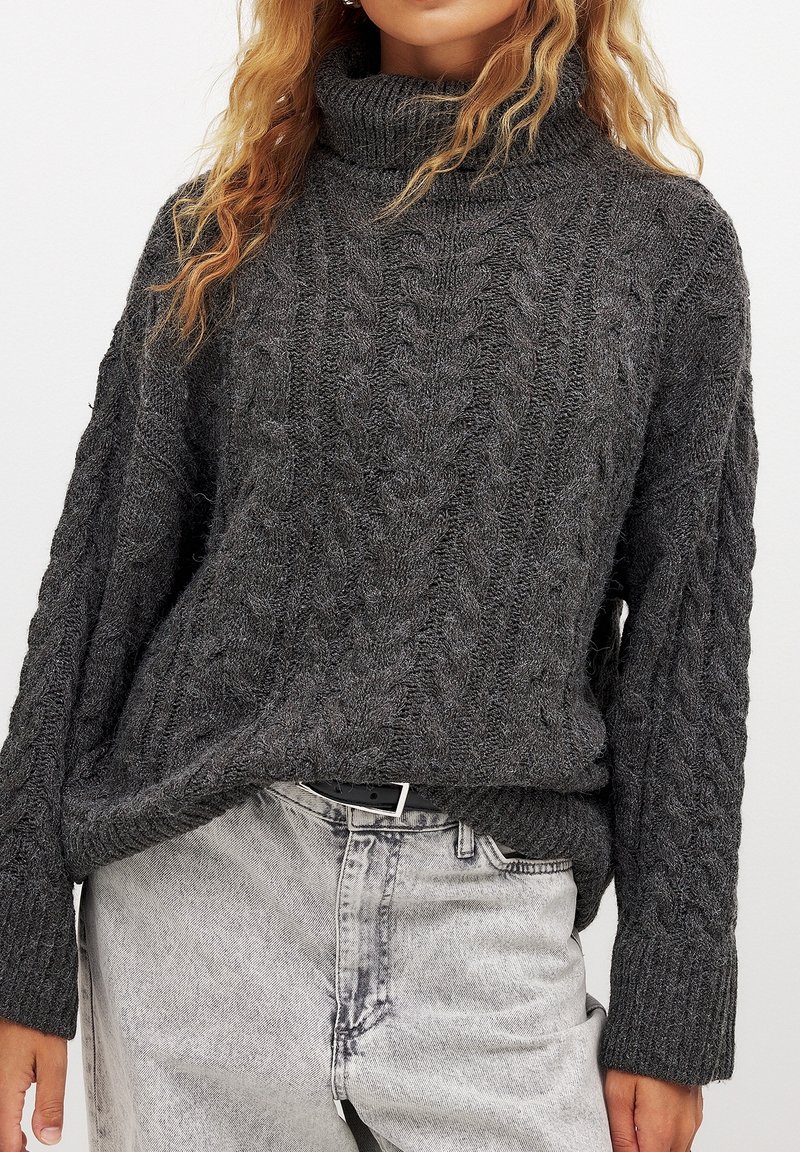 Dunkelgrauer Kabelstrickpullover mit hohem Rollkragen, weiten Ärmeln und strukturiertem Stoff. Kombiniert mit hellgrauen hochgeschnittenen Jeans.