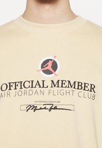Chemise beige avec le logo Air Jordan Flight Club, une silhouette rouge de Jumpman, le texte « Official Member » et une signature imprimée en dessous.