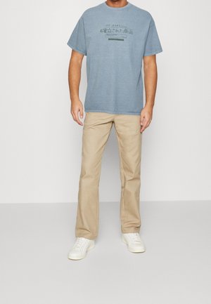 Modrá bavlněná trička s grafickým potiskem, kombinovaná s béžovými chinos kalhotami a bílými teniskami. Jednoduchý design, pohodlný střih a jednobarevné provedení.