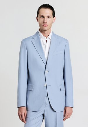Uomo con capelli slicked-back, che indossa un completo azzurro chiaro e una camicia bianca, in piedi contro uno sfondo grigio chiaro uniforme.