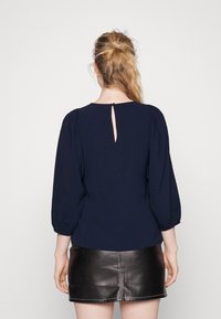 Maglione blu navy con maniche a sbuffo, scollo rotondo e dettagli a chiave sulla schiena. Tessuto morbido, adatto per un look casual.