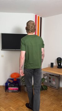 Groen T-shirt met korte mouwen, slim fit, gecombineerd met donkergrijze spijkerbroek. Moderne inrichting met een kleurrijke muur, tv en diverse meubelstukken.