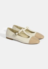T-Strap Ballerinas aus cremefarbenem Leder mit einer glänzenden, beigen Kappe. Mit einem Schnallenriemen, niedrigem Absatz und glatter Textur.