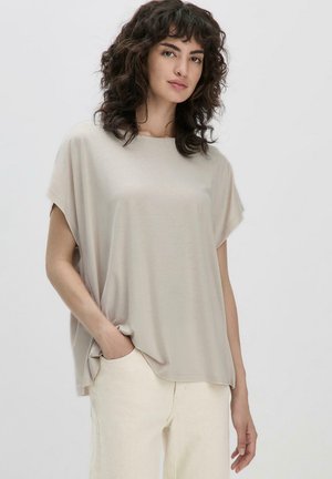 Top beige morbido e largo con maniche corte, scollatura rotonda e texture soffice, abbinato a pantaloni chiari. Design minimalista, senza fantasie.