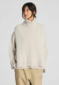 Mujer de cabello oscuro con un suéter beige de cuello alto y holgado y pantalones marrón claro, de pie con las manos en los bolsillos frente a un fondo sencillo.