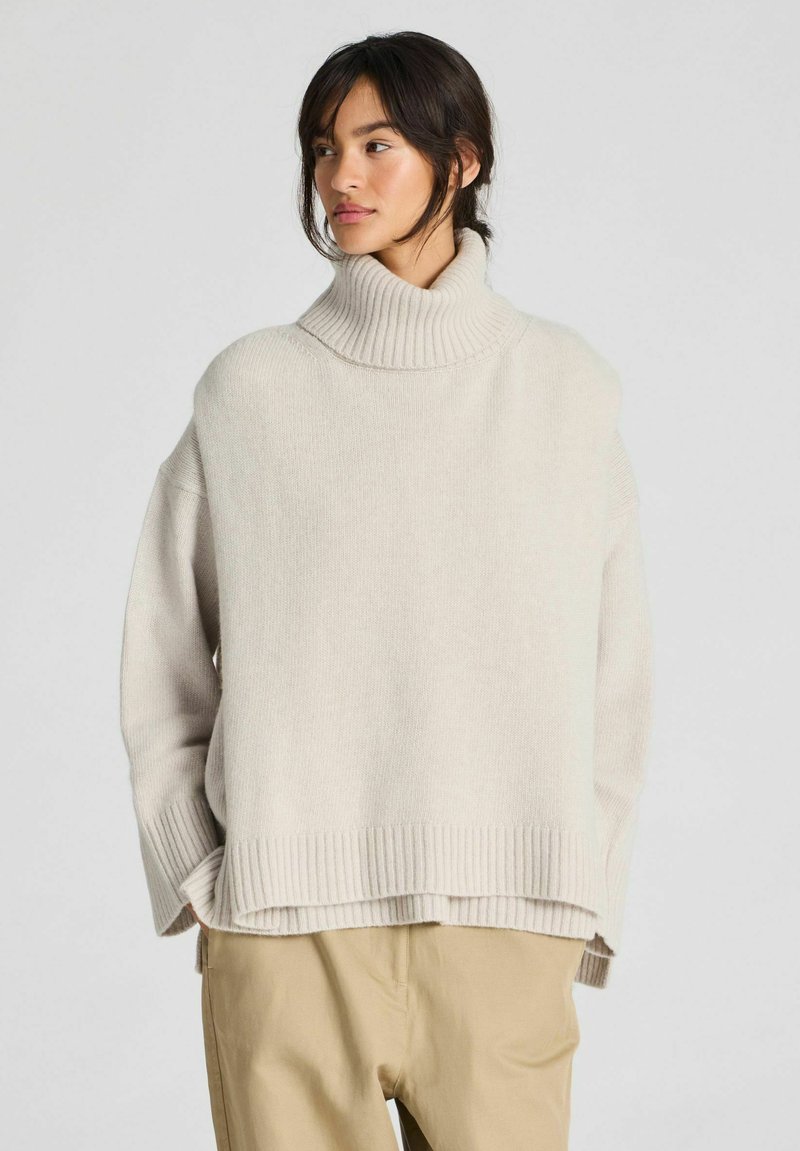 Mujer de cabello oscuro con un suéter beige de cuello alto y holgado y pantalones marrón claro, de pie con las manos en los bolsillos frente a un fondo sencillo.