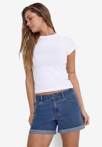 Λευκό κοντομάνικο t-shirt σε εφαρμοστή γραμμή, συνδυασμένο με μπλε τζιν σορτς που έχουν διπλωμένα μανίκια και μπροστινές τσέπες, μέσα σε στούντιο.