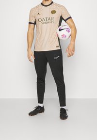 Béžový fotbalový dres s texturovaným vzorem, černými akcenty a logem "QATAR AIRWAYS". Černé sportovní kalhoty s logem Nike. Hráč držící fotbal.