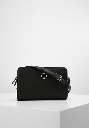 Sort rektangulær crossbody taske med justerbar strop og et lille rundt metal logo foran, placeret på en hvid piedestal.