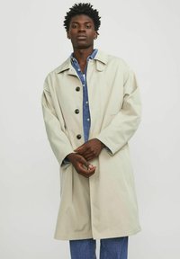 Beige regnrock med avslappnad passform, stor krage, svarta knappar och sidofickor, bärs över en denimskjorta, visad av en modell.