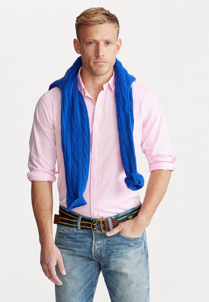Suéter de punto trenzado azul brillante sobre una camisa de cuadros rosa claro, combinado con jeans oscuros y un cinturón multicolor con una hebilla de latón.
