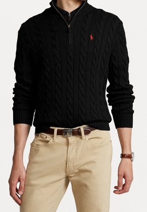 Mann trägt schwarzen Strickpullover mit Viertelreißverschluss und rotem Logo, beige Hose, braunen Ledergürtel und braune Lederarmbanduhr.