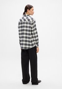 Camisa de franela a cuadros en blanco y negro con cuello abotonado y mangas largas, combinada con pantalones sueltos negros y zapatos oscuros.