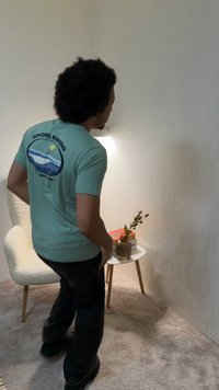 Homme aux cheveux bouclés portant un t-shirt bleu « Ripcurl Surfing » debout près d'une petite table blanche avec des livres et un vase dans une pièce d'angle avec moquette.