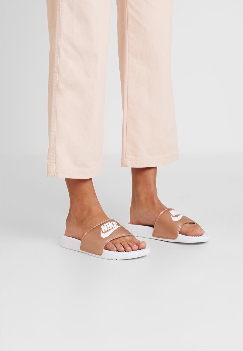 Pieds portant des sandales Nike slide beiges avec des semelles blanches, associées à un pantalon court rose clair sur un fond uni.