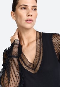 Zwarte top met doorschijnende stippenkant lange mouwen en een diepe V-hals, met textuurstof en gerimpelde accenten voor extra detail.