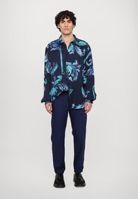 Marineblauw shirt met lange mouwen met levendige turquoise bloemenpatronen. Gecombineerd met donkermarineblauwe broeken en zwarte veterschoenen.