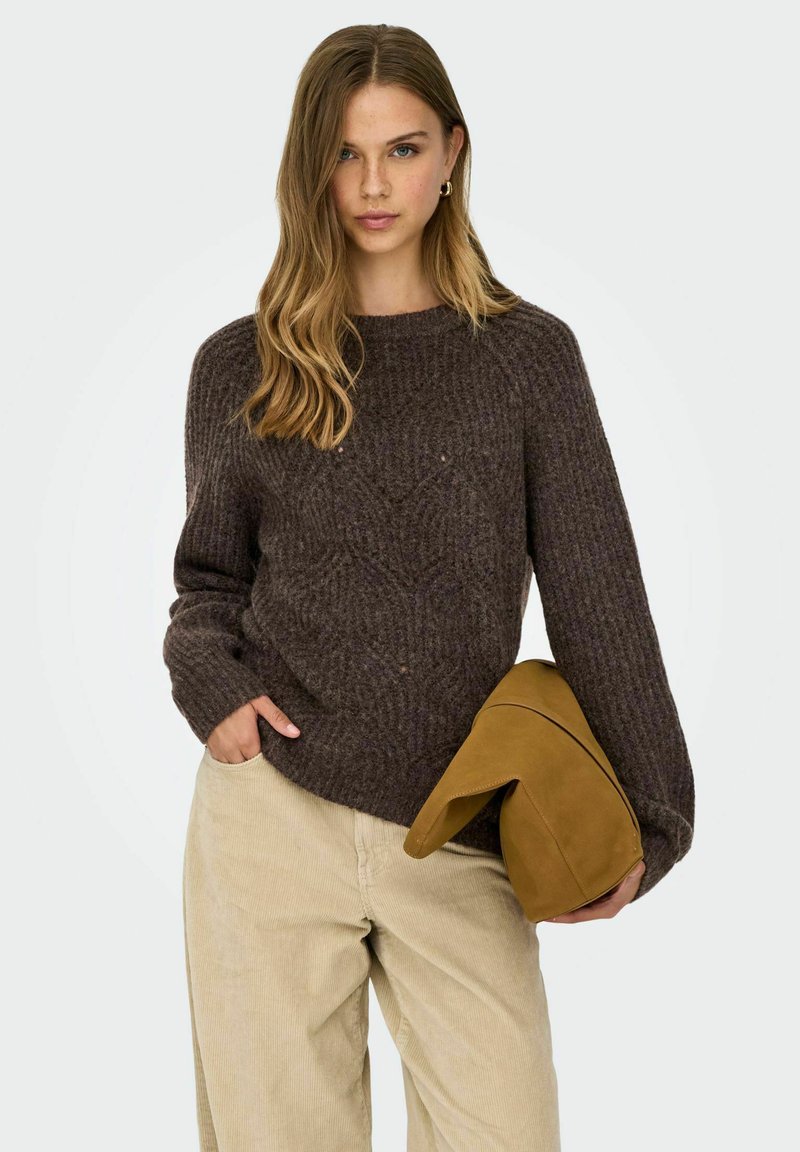 Bruine geribbelde gebreide trui met een ronde halslijn, gecombineerd met beige corduroy broek. Het model houdt een lichtbruine suede tas met een gevouwen klep vast.