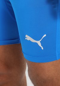 Blå atletiske shorts lavet af glat, strækbart stof. Har et hvidt Puma-logo på venstre lår. Tætsiddende design med syede sømme.