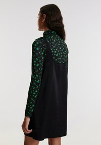 Zwarte mouwloze jurk over een langmouwen top met een groen bloemenpatroon. Glad stof en een getailleerde taille. De achterkant benadrukt het ontwerp.
