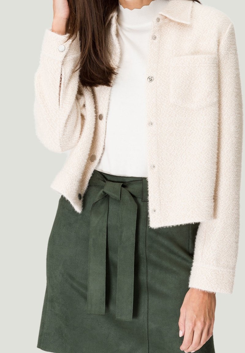 Mujer vistiendo una chaqueta abotonada de textura cremosa sobre una blusa blanca, combinada con una falda verde oscuro de cintura alta atada con un cinturón de tela.