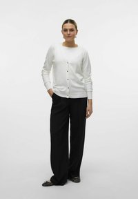 Cardigan bianco con scollatura a V e bottoni dorati, abbinato a pantaloni neri larghi. Il tessuto appare liscio, creando un look pulito e minimal.