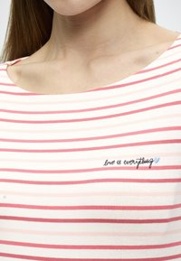 Ριγέ T-shirt με απαλό ύφασμα· διαθέτει οριζόντιες ρίγες σε ανοιχτό ροζ και κόκκινο, στρογγυλή λαιμόκοψη και κεντημένο κείμενο: «η αγάπη είναι τα πάντα» με μια ανοιχτό μπλε καρδιά.