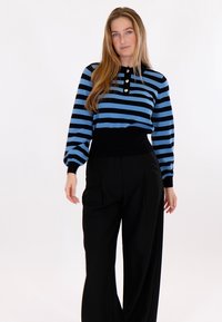 Žena s dlouhými vlasy, která nosí modrý a černý pruhovaný svetr s dekorativními knoflíky a černé široké kalhoty s vysokým pasem na bílém pozadí.