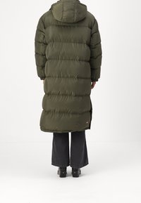 Lång, olivgrön puffjacka med huva, med ett kviltat mönster, något utställd vid fållen, kombinerad med grå utställda jeans och mörka skor.