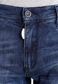 Denim jeans in dark blue with a textured finish. Features include a metal button at the waist and a front pocket with visible stitching.