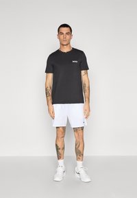 Svart t-skjorte med korte ermer som har hvit "BOSS" logo, sammen med hvite shorts og hvite joggesko. Tatauer synlige på armer og ben.