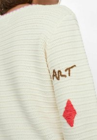 Trikotaža u boji šampanjca s teksturiranim prugama. Sadrži smeđu vezenu oznaku "ART" i crvenu dijamantnu aplikaciju na rukavu.
