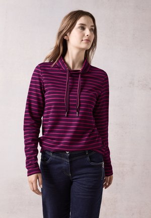 Jersey de manga larga en color púrpura oscuro con atrevidas rayas horizontales rosas, con cuello tipo cowl y cordones. Combinado con jeans oscuros.