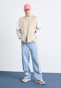 Gilet trapuntato beige sopra una felpa grigia, abbinato a pantaloni della tuta azzurro chiaro e sneakers beige. Gli accessori includono un berretto da baseball rosa.