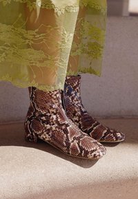 Bruna och krämfärgade ankellånga stövlar med ormskinnsmönster, en slät, glansig yta och låg blockklack, kombinerade med ett genomskinligt grönt lager med spetskant.