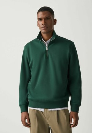 Uomo che indossa un pullover verde scuro con zip a quarto sopra una camicia chiara e pantaloni beige, in piedi contro uno sfondo chiaro uniforme.