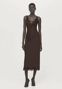 Bruine slipjurk met een kanten rand bij de halslijn en zoom, met een rechte silhouet. Materiaal lijkt glad en lichtgewicht.