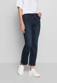 Granatowe dżinsy o prostym kroju, z kontrastowym przeszyciem i pięcioma kieszeniami, noszone z białymi trampkami.