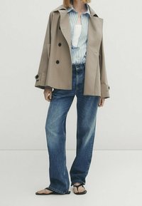 Henkilö, joka pukeutunut beigeen kaksiriviseen takkiin sinisävyisen raidallisen paidan päällä, leveälahkeisiin farkkuihin ja mustiin varvastossuihin, seisoo yksinkertaisen taustan edessä.