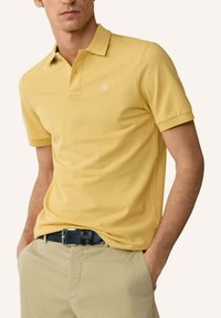 Homme portant un polo jaune à manches courtes rentré dans un pantalon beige avec une ceinture noire, les mains dans les poches, sur un fond clair uni.