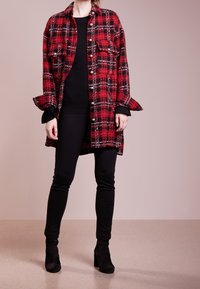 Röd och svart rutiga oversize-jacka med två bröstfickor, metallknappar och uppvikta ärmar, bärs över en svart topp och leggings.