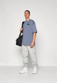 Pantalones deportivos cargo grises, camiseta azul con logo verde y zapatillas blancas. Modelo sosteniendo una bolsa negra de hombro, posando frente a un fondo neutro.