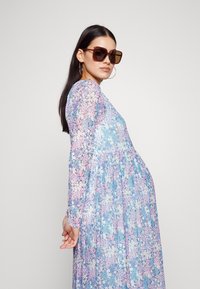 Blumenmotiv Kleid mit langen Ärmeln in Blau-, Rosa- und Grüntönen, mit einer gerafften Taille und fließendem Design. Das Model trägt große Sonnenbrillen.