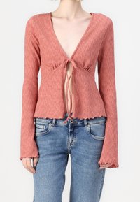 Kvinna som bär en texturerad, långärmad, kort pink topp med knytning fram, ihop med ljusblå denimjeans.