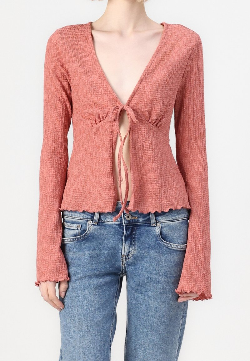 Kvinna som bär en texturerad, långärmad, kort pink topp med knytning fram, ihop med ljusblå denimjeans.